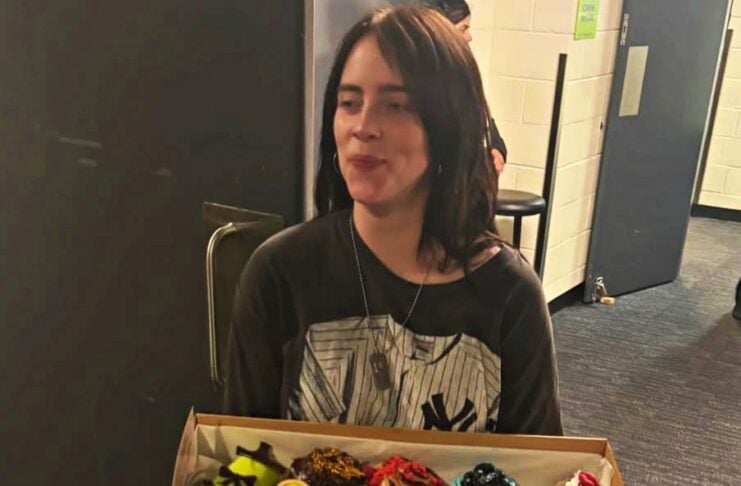 Young woman in a dark shirt holds a cardboard box of assorted donuts in a hallway.