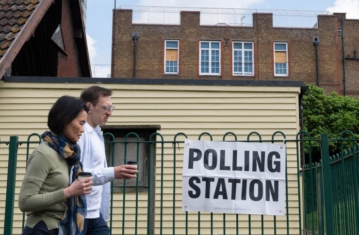 Two people walk past a polling station sign on a green fence, each holding a cup of coffee.