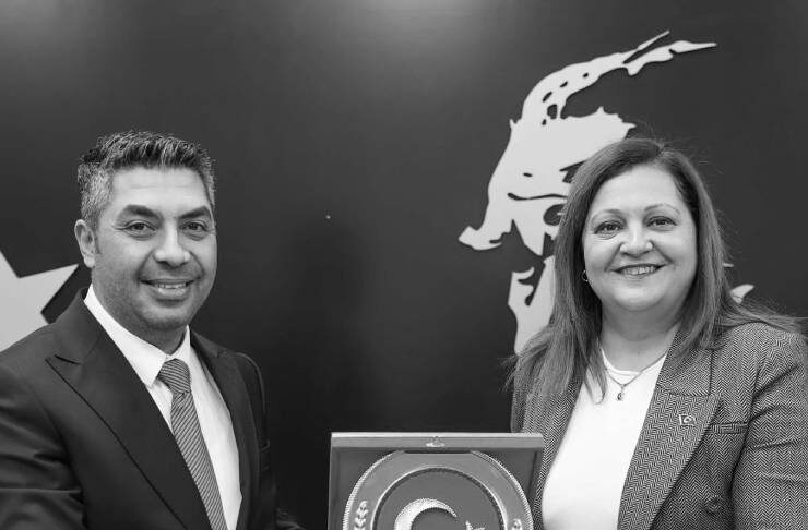 Two professionals in business attire present a plaque featuring a crescent moon and star emblem. Behind them, a dark backdrop with a large abstract figure.