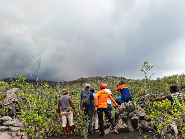 Rescue workers in orange jackets and helmets stand on volcanic rock, looking toward a dark smoke plume in the cloudy sky.