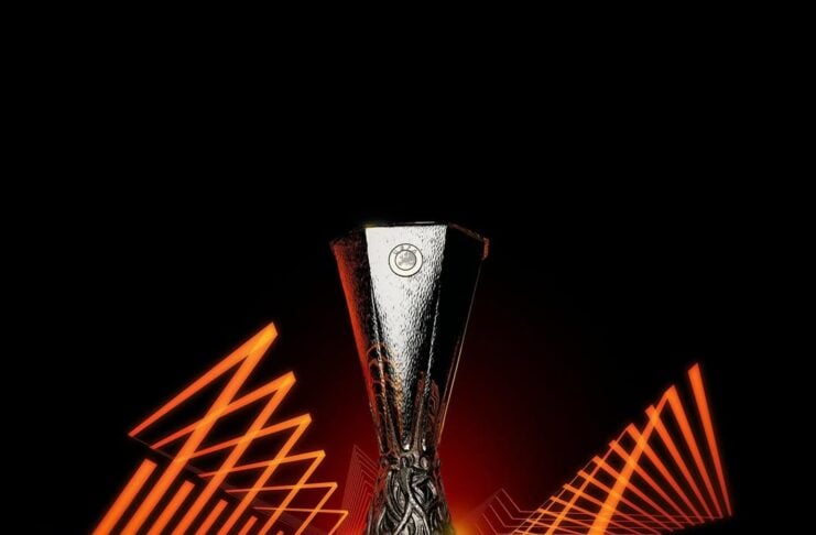 A silver UEFA Europa Conference League trophy centered on a black background with orange neon angular lines framing it.