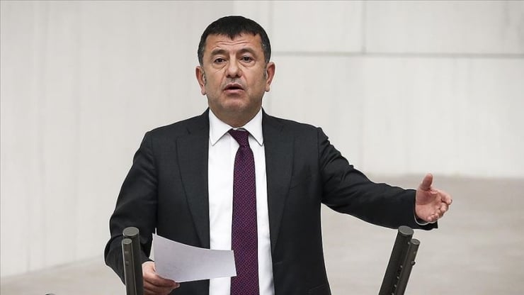 Man in a dark suit and purple tie speaks, holding a sheet of paper and gesturing with his right hand in a formal setting