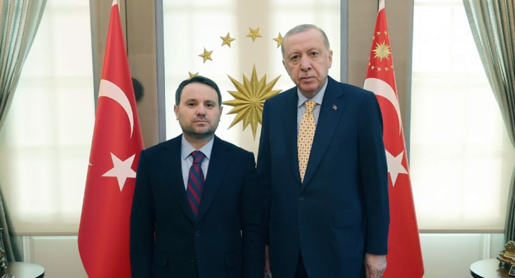 Two men in suits pose for a formal photo in front of Turkish flags and a gold emblem behind them