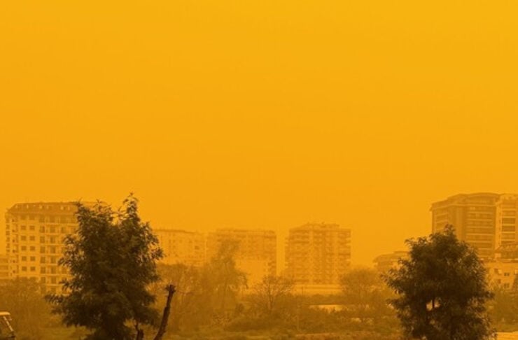 Urban street scene with parked cars and a person walking, trees in the foreground and greenhouses in the midground, tall buildings in the hazy orange sky/polluted air.