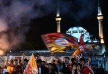 Nighttime crowd celebrating in a street, waving flags and filming with phones, with smoke in the air and a lit mosque with minarets in the background.