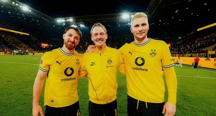 Three Borussia Dortmund players in yellow home jerseys posing together on the football pitch at night in a filled stadium.