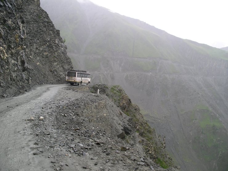 Bus parked on a narrow rocky cliff road with steep drop-offs on the right and mountains in the background