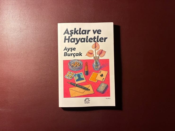 Book cover: Aşklar ve Hayaletler by Ayşe Burçak, with a colorful illustration of a plant, cards, lipstick, pen, and envelopes on a pink rectangle on white background.