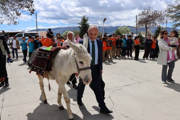A suited man leads a white donkey with a decorative saddle as a crowd watches at an outdoor event.