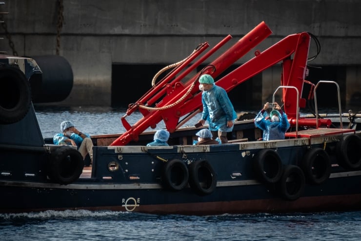 Crew in blue protective suits and helmets stand on a dark blue workboat, next to a large red crane on deck by the water.