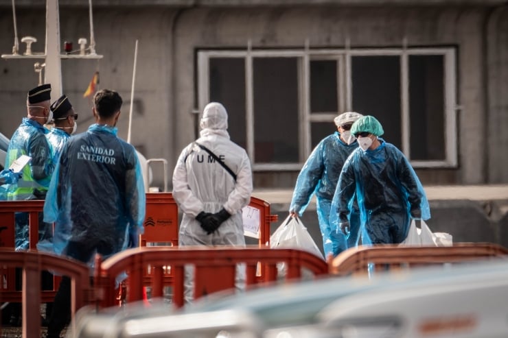 Medical responders in blue PPE and hazmat suits carry white bags outside a building near orange barriers.