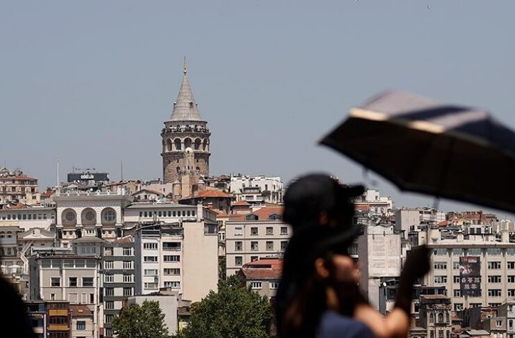 Istanbul cityscape with the Galata Tower rising above hillside buildings, seen from a waterfront promenade with blurred people in the foreground.