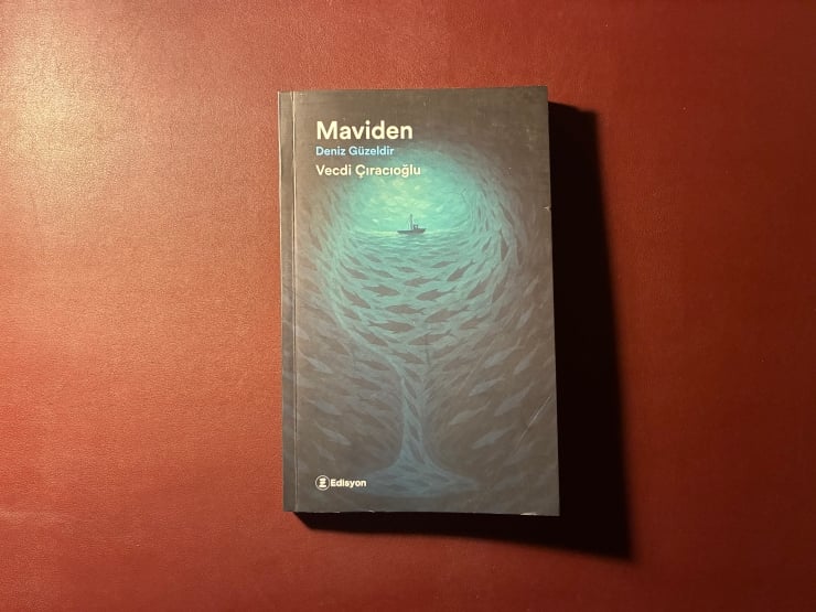 Dark book cover of 'Maviden' with a teal concentric wave design and a small boat at center, on a red tabletop (authors: Deniz Güzeldir, Vecdi Çiraçoğlu).