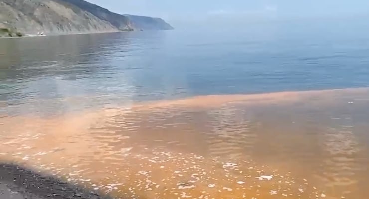 Shoreline with calm blue water and orange-brown tide pool along the beach, cliffs in the distance on the left.