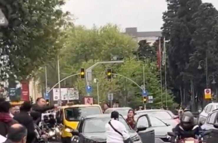 Crowded city intersection with pedestrians in the street and multiple vehicles, including a motorcycle approaching from the right.