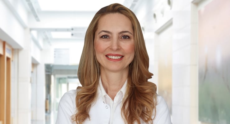 Smiling professional woman with long wavy hair in a white blouse standing in a bright office hallway.