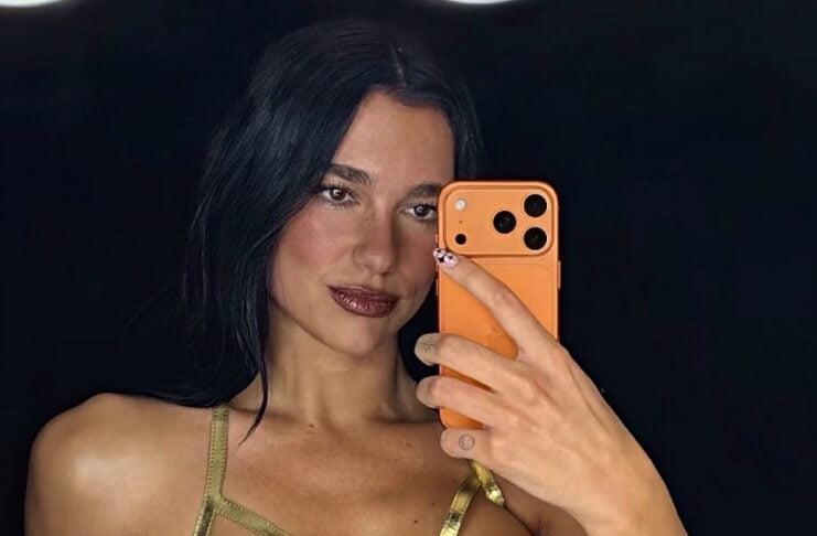 Woman taking a mirror selfie in a metallic gold corset top, holding an orange smartphone against a dark background.
