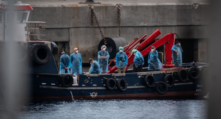 Crew in blue disposable coveralls and masks stand on a dark blue workboat with a red crane, near a concrete dock beside calm water.