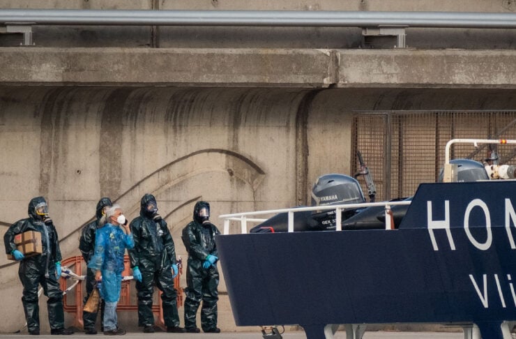 Group of people in hazmat suits standing on a dock beside a blue boat, one person holding a box in the background.