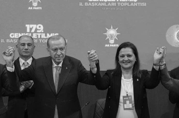 Black-and-white photo of four suited individuals on a stage, holding hands raised in unity, with a woman in a white blouse wearing a badge, at a political event backdrop with party logos.