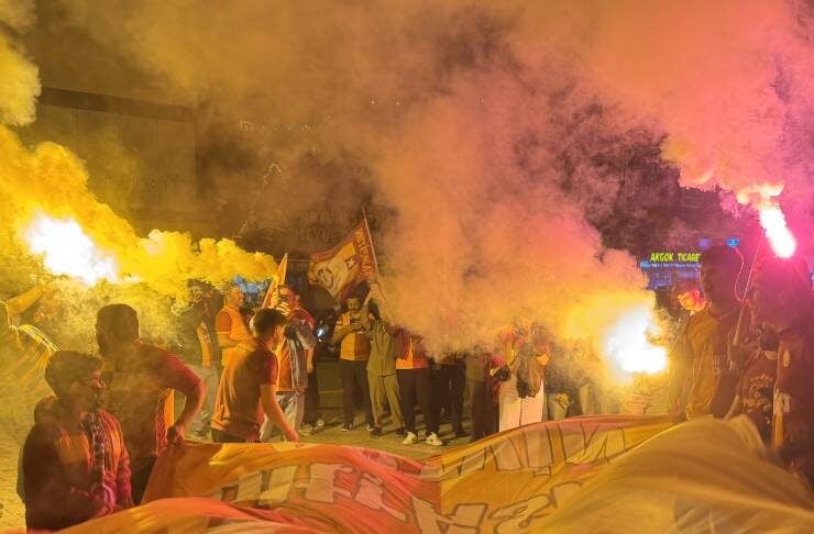 Crowd holding a large banner during a night protest with orange smoke from flares surrounding them, flags visible in the background.