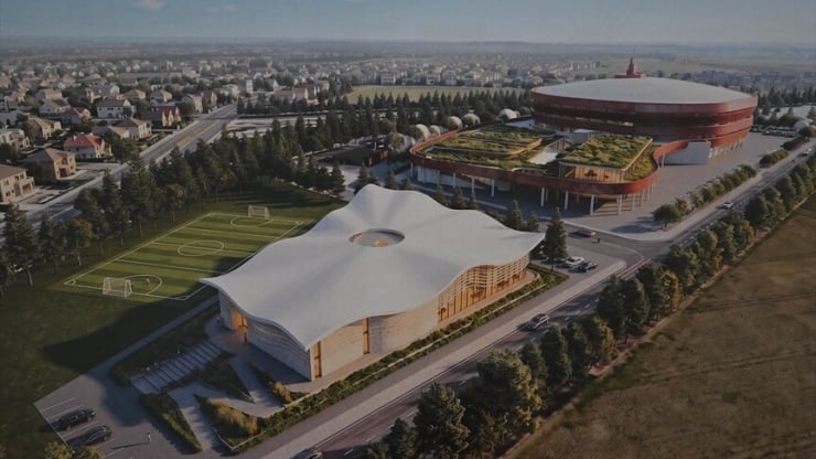 Aerial view of a modern sports complex with a white tent-like roof building beside a circular arena, surrounded by fields, trees, and a residential area.