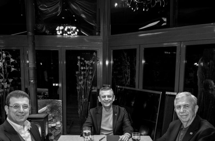 Three men sit at a rectangular table in a dimly lit restaurant, each with a glass of wine, suggesting a business dinner.