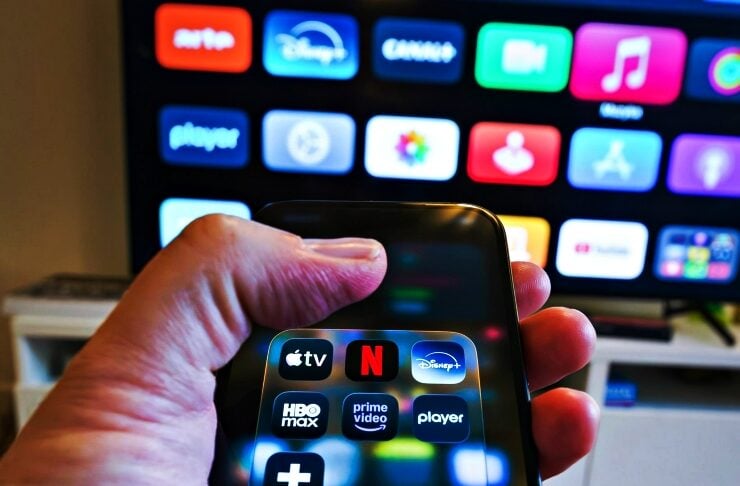 Hand holding a smartphone showing a folder of streaming apps (Netflix, Disney+, HBO Max, Prime Video, YouTube) with a TV apps screen in the background
