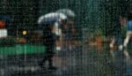 Rainy urban scene viewed through a rain-speckled window; blurry figures with an umbrella in the background.
