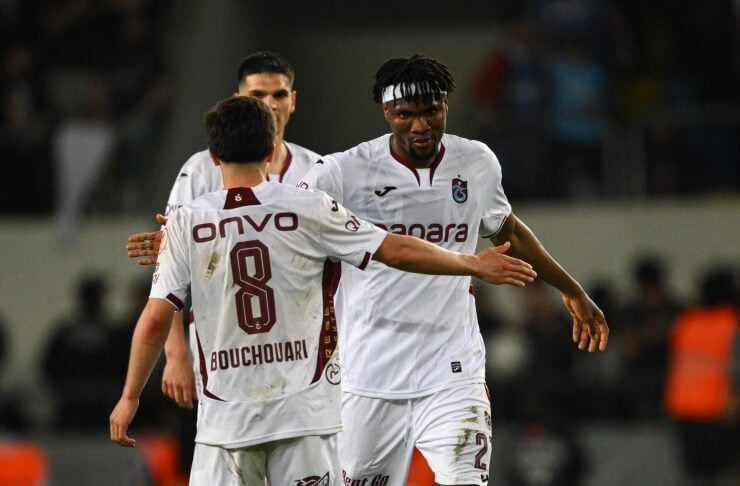Two soccer players in white uniforms celebrate on the field, arms extended in a congratulatory high-five after a goal.