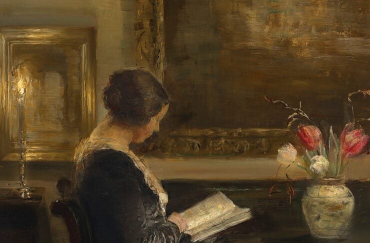 Woman seated at a dark desk reading a book, with a vase of red and white flowers nearby