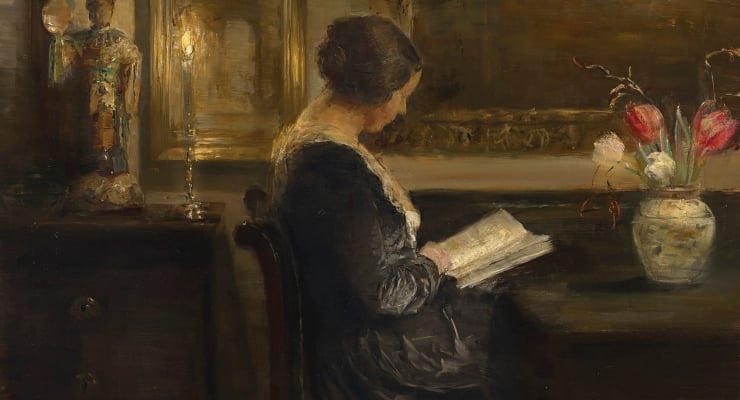Woman in a dark dress sits at a table reading a book in a softly lit interior, with a vase of flowers nearby.