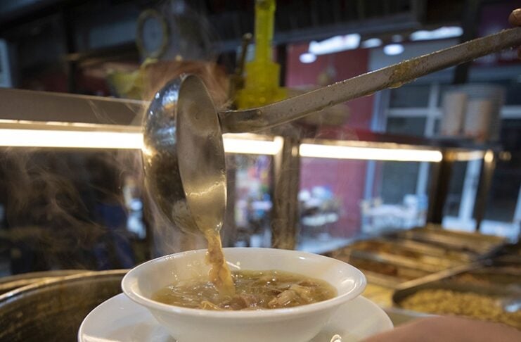 Bowl of hot soup being ladled from a large pot with steam rising in a cafe or market setting.