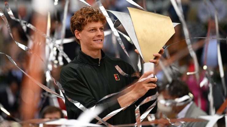 Man in a black jacket holds a large gold trophy as silver streamers swirl around him at a celebration event.