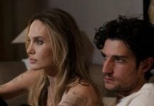 A blonde woman with visible tattoos sits on a couch next to a man with dark hair; both gaze toward something off-camera.