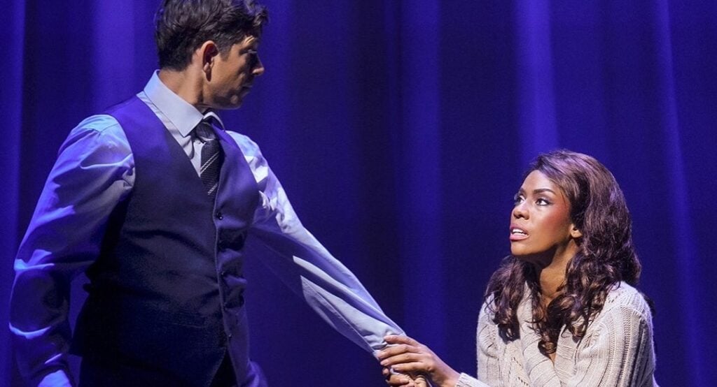 Dramatic stage moment: a man in a blue vest extends his hand to a woman who is seated or kneeling, under blue theater lighting.