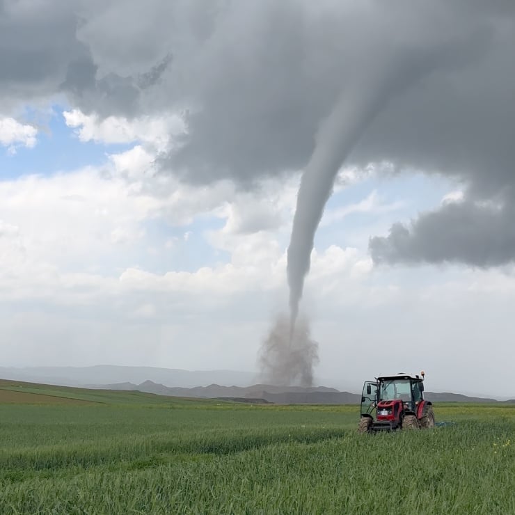 Red tractor in a green field as a dust-tornado spins up from the ground under a stormy sky.