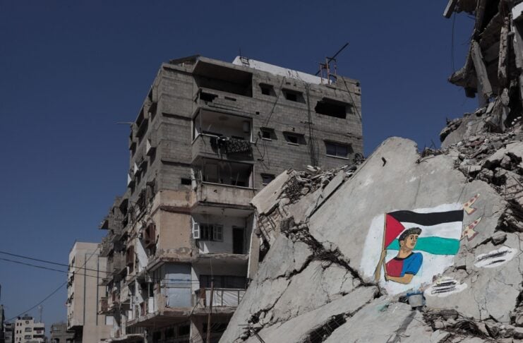 Ruined city street with a partially collapsed building, rubble on the ground, and a mural of a person holding a Palestinian flag on a cracked wall.