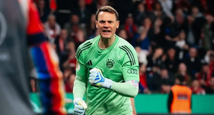 Goalkeeper in a bright green kit and white gloves shouts on the field during a match with a crowded stands backdrop.