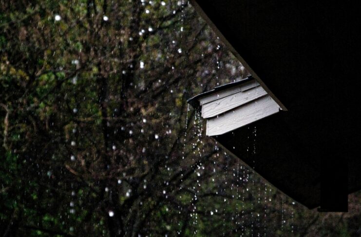 Rain falls from the edge of a house roof overhang, with a dark yard and trees in the background.