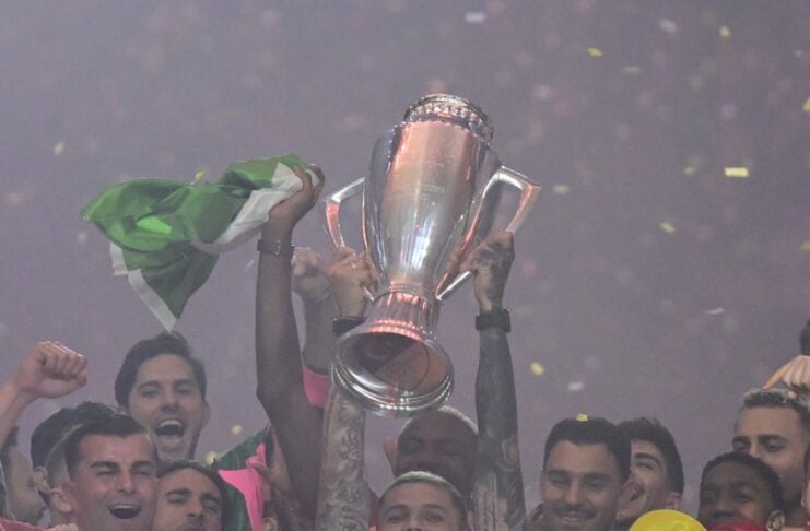 Soccer team celebrates victory, lifting a large trophy amid confetti and cheering players in orange jerseys.