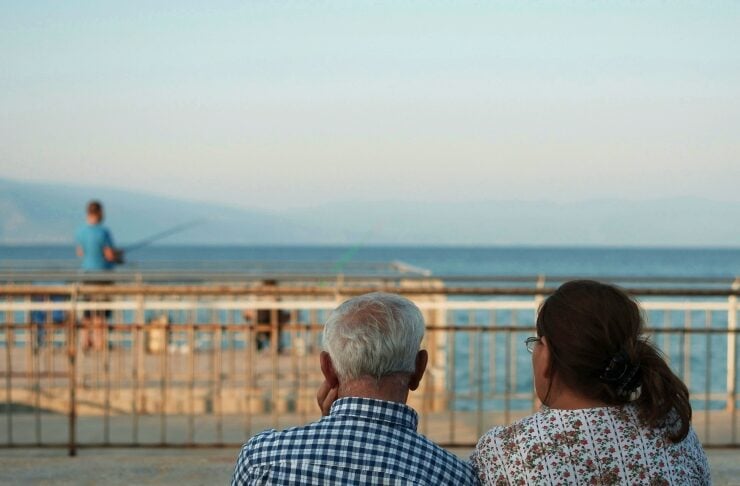 Two seniors sit on a wooden bench facing the sea, watching a person fishing on a pier. In the distance, blue water and mountains.