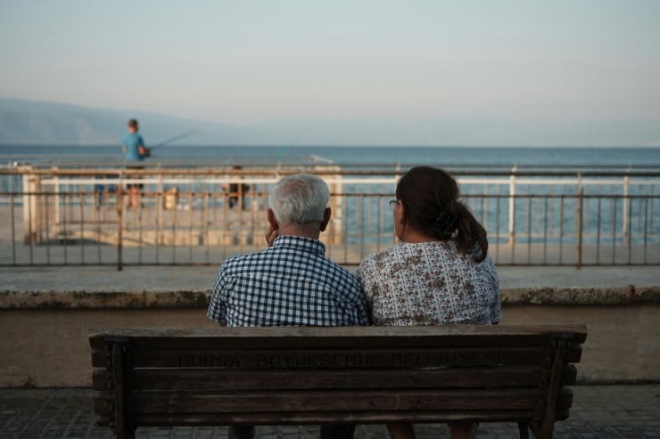 Elderly couple sitting on a wooden bench by the seaside, facing the ocean with a fisherman in the background.