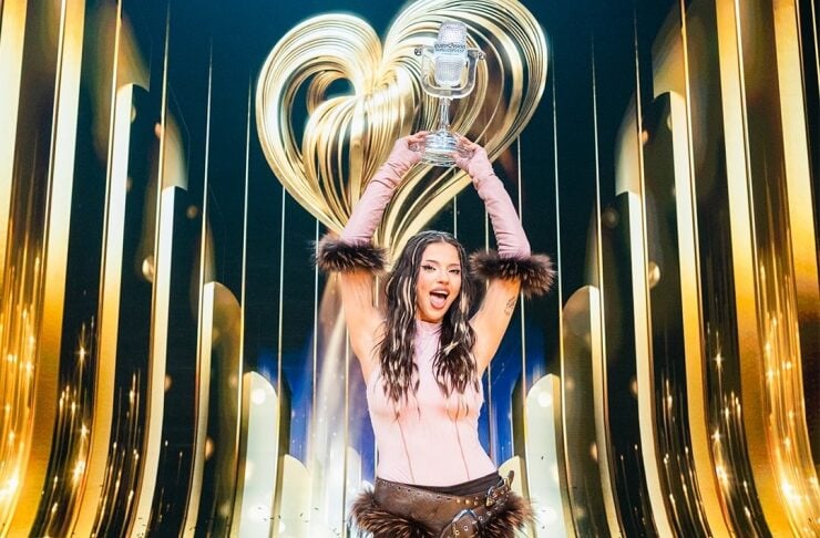 Woman in pink top and fur-trim shorts raising a trophy on a shiny stage with gold decor.