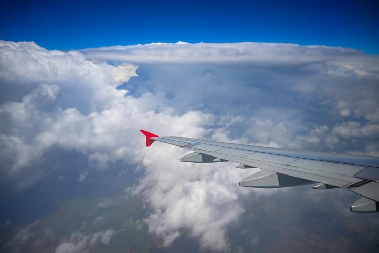 Airplane wing with a red wingtip slicing through a sea of white clouds under a deep blue sky (in-flight view).