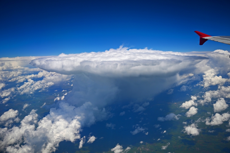 View from an airplane window: red-tipped wing on the right, vast blue sky and a towering cloud formation over the land below.