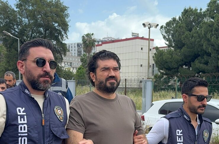 Two officers in SIBER vests escort a man in a brown T‑shirt outdoors near a fence.