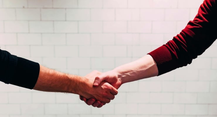 Two people are shaking hands in front of a pale brick wall, signaling an agreement or partnership, close-up of hands and forearms touched by light.