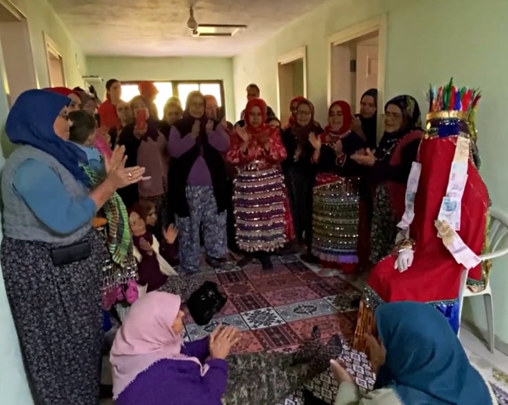 Group of women in bright, traditional dresses clapping around a seated performer in a red robe with a tall feathered headdress in a festive room./