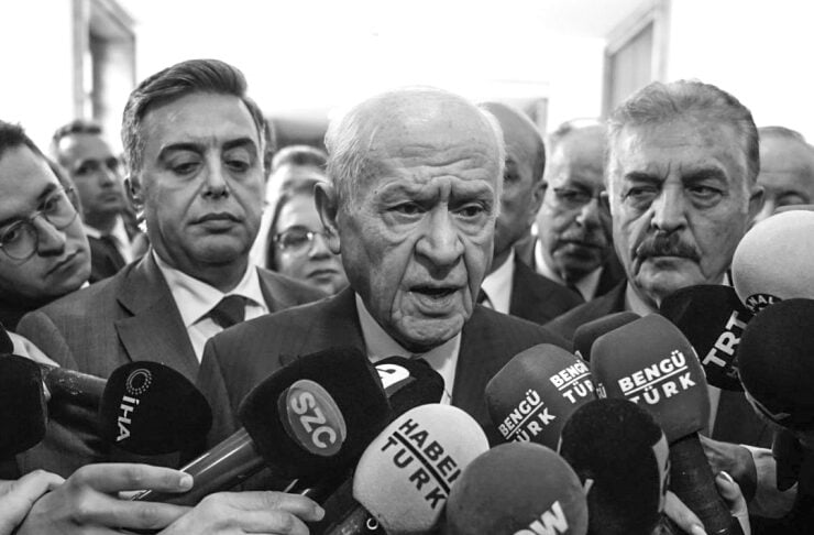 Older man in a suit speaks to a cluster of reporters holding microphones at a press conference in black and white.